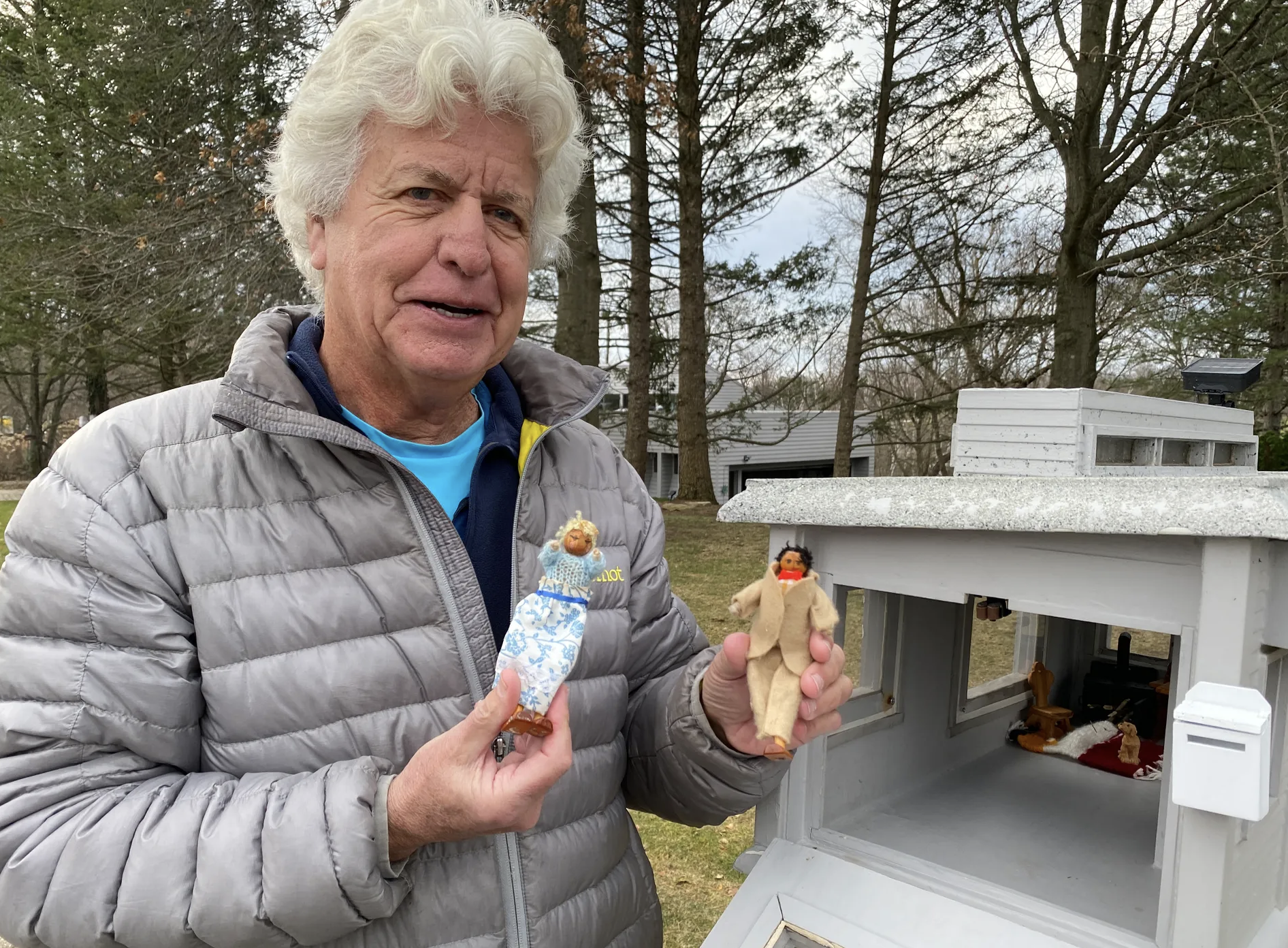 Women in a coat holding two dolls in her hands with a house mailbox.