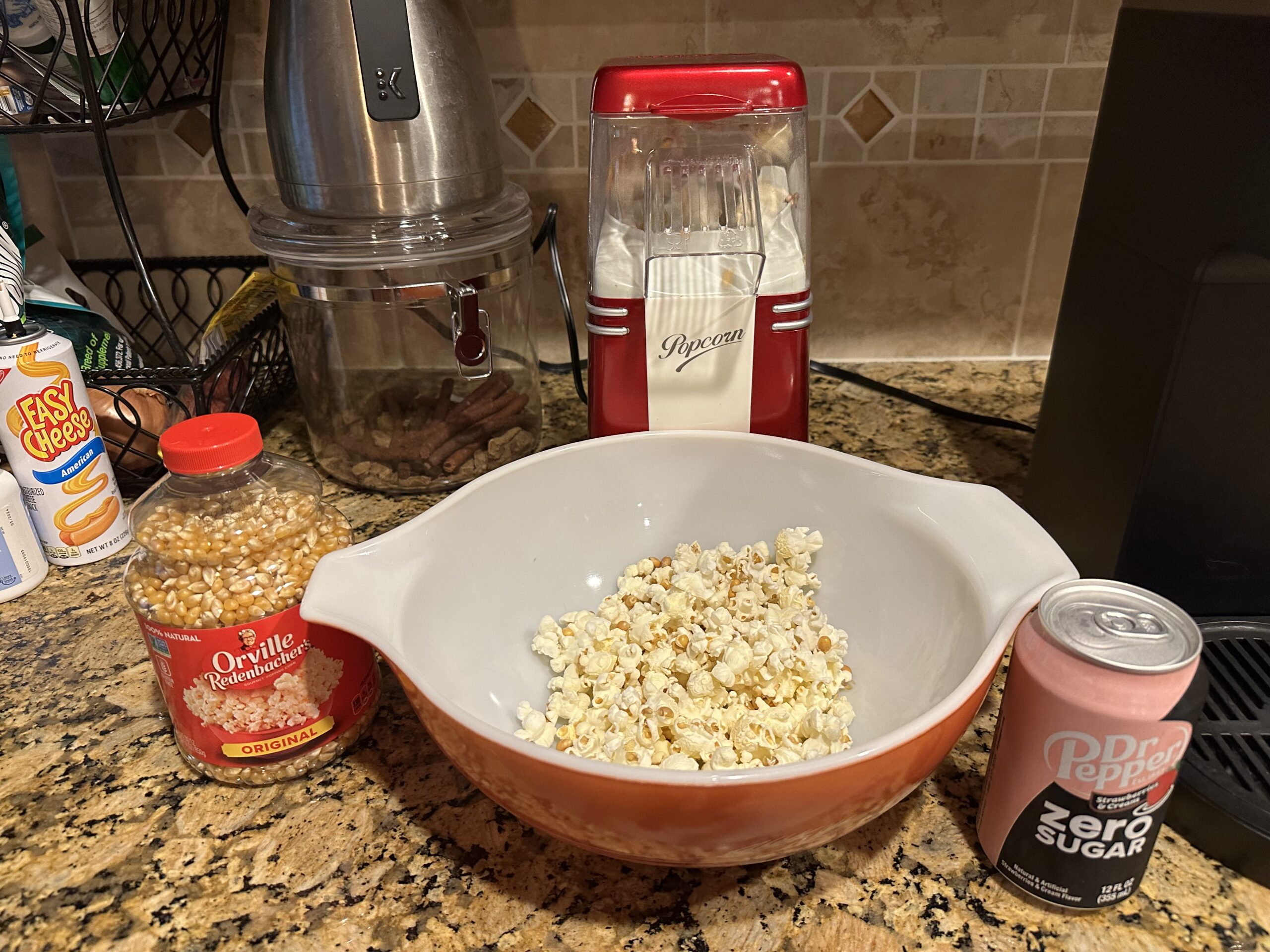 Bowl of popcorn, popping corn in container, and Dr. Pepper zero sugar sitting on the counter.